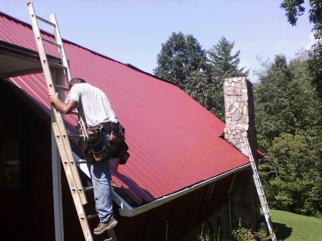 Roofer on a ladder beside a red metal roof and stone chimney — Soffit & Fascia Repair by Roofing Brothers in Bloomdale